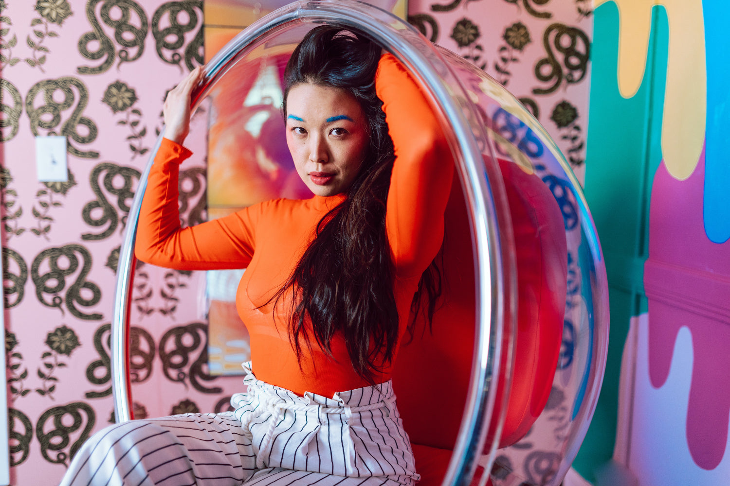 Woman in vibrant clothes and makeup poses in a large acrylic egg chair