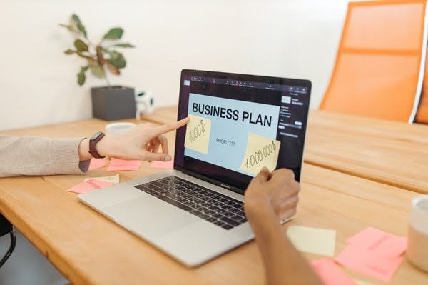 An open laptop with BUSINESS PLAN on screen and fingers pointing to notes.