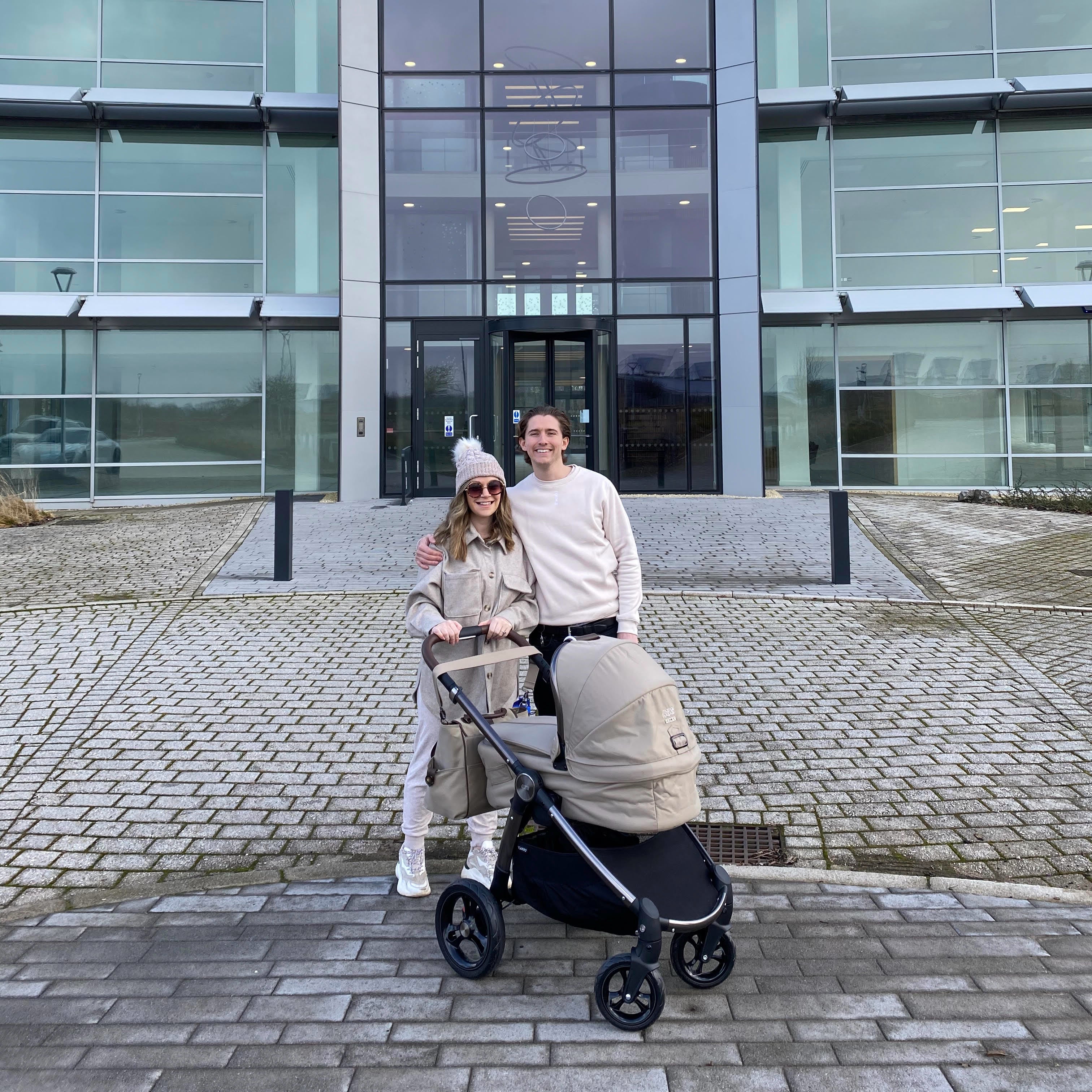 Dan and Mel Marsden wearing beige clothing along with their daughter in a stroller backdropped by their new headquarters. 