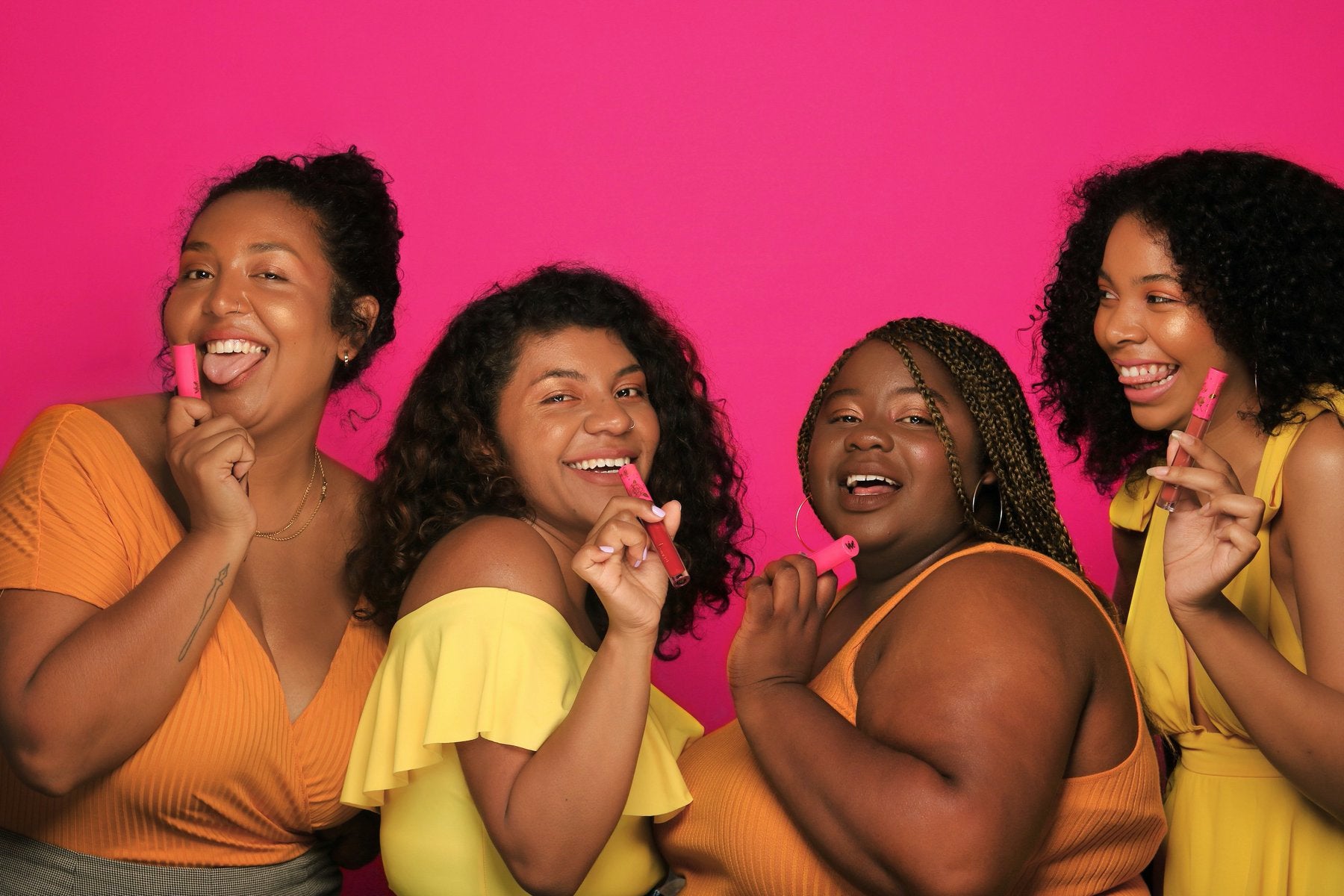 Four women wearing yellow hold lipsticks against a pink background