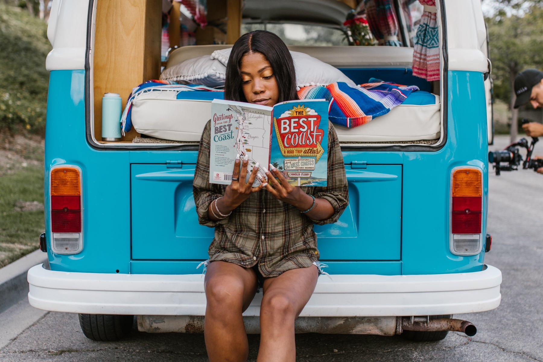 A woman sits on the rear bumper of a camper van and reads a book about travel