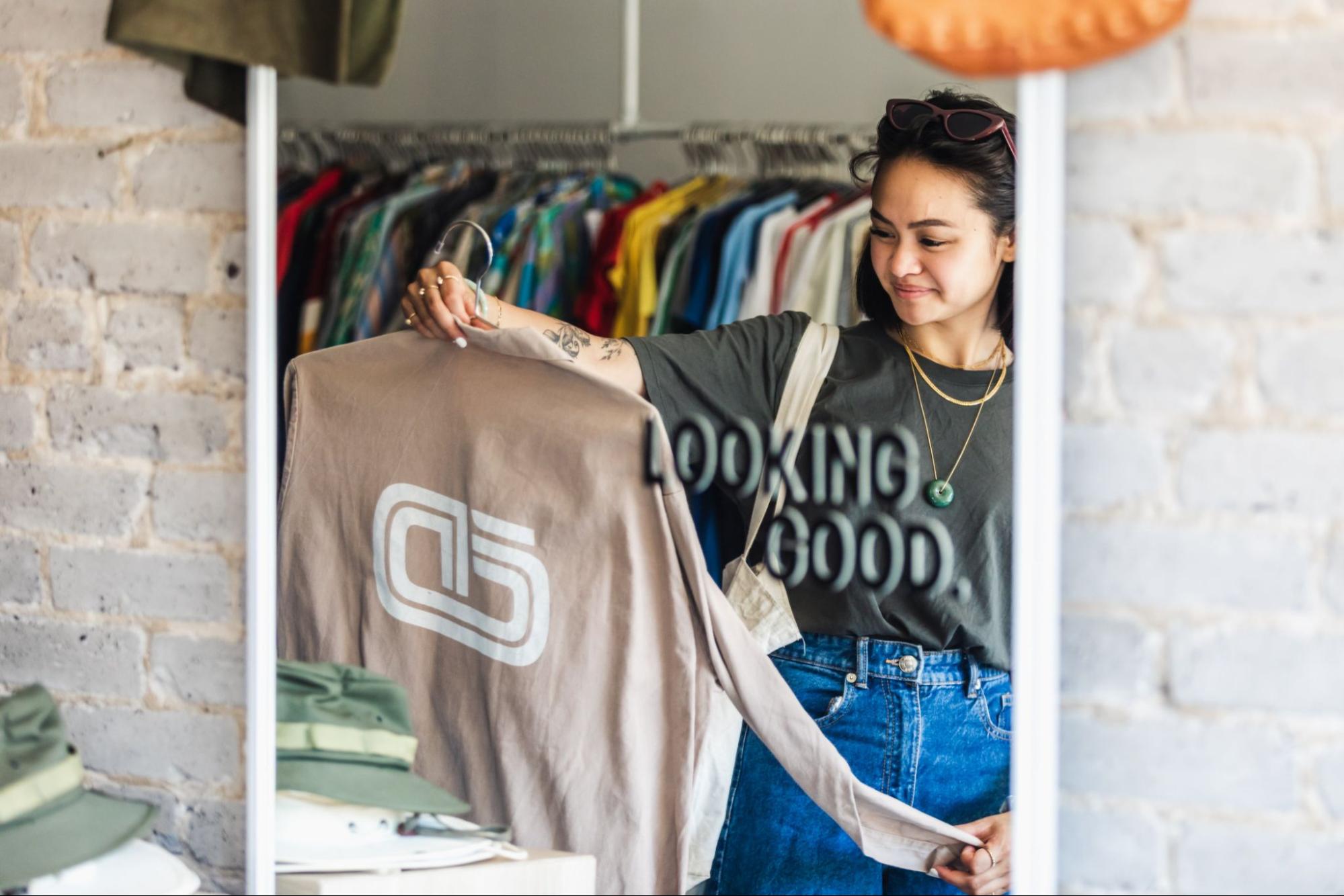 A young woman holding up a top in a clothing store, seen through a mirror.