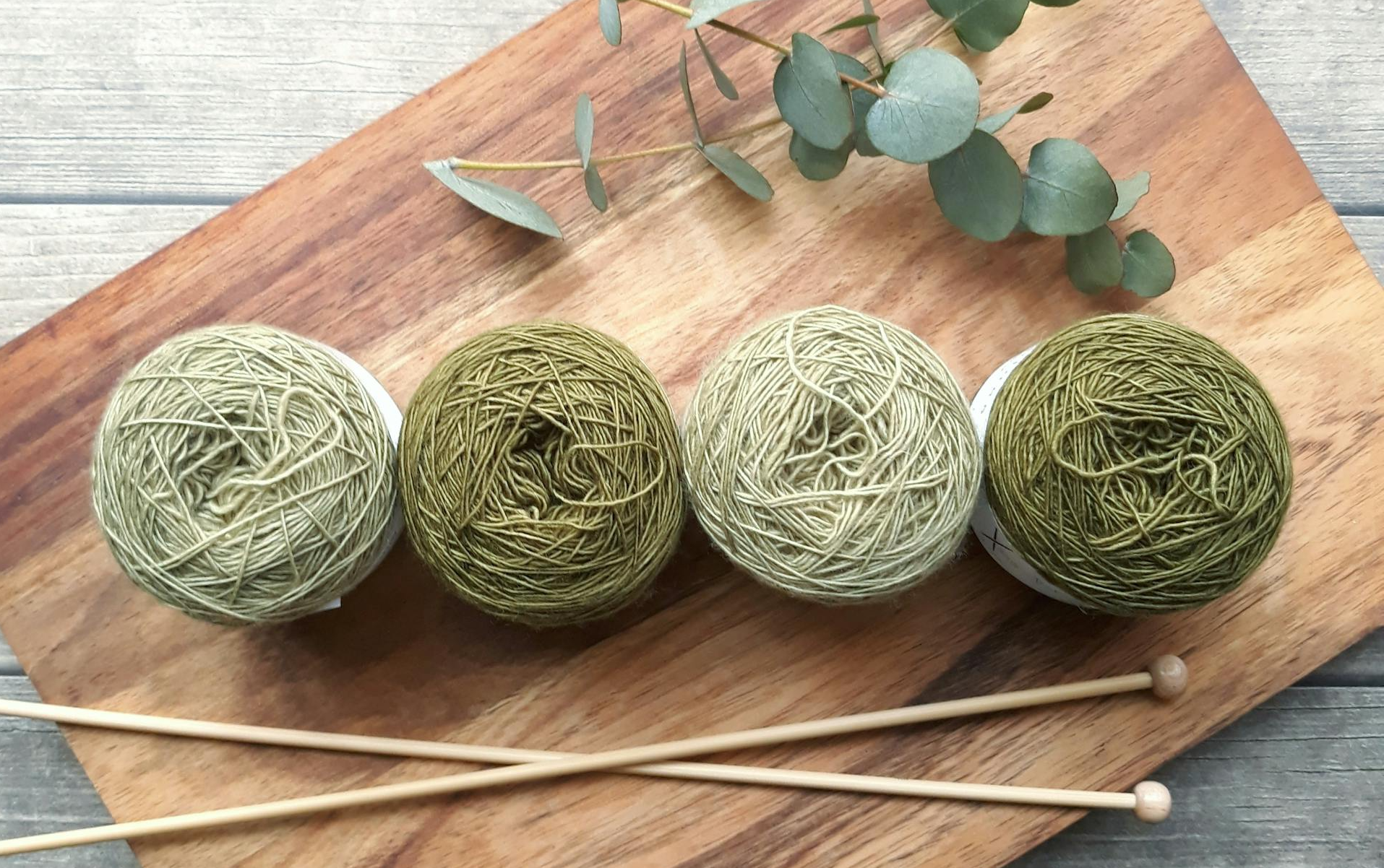 Four balls of yarn lined up above a pair of knitting needles on a wooden cutting board.