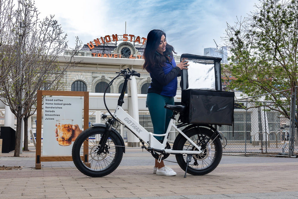 A woman unloading food from a delivery bag on the GOTRAX F2 folding electric bike with wide tires and a rear storage rack.