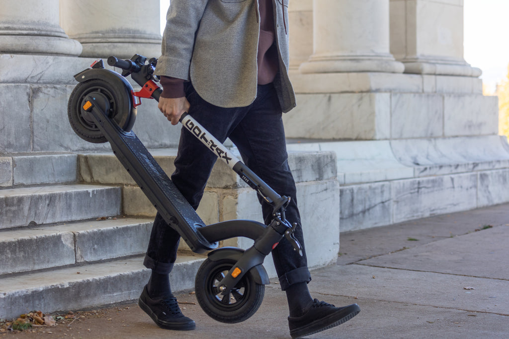 A man carrying the GOTRAX G4 folding electric scooter with a digital display down a set of stairs.