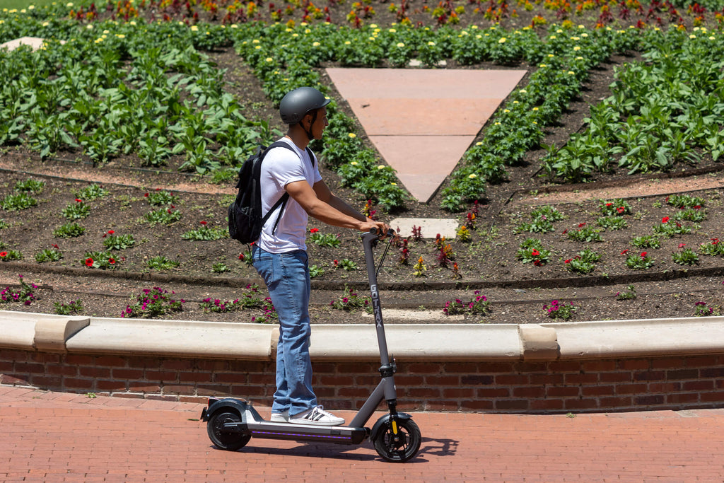 A young man riding the GOTRAX G5 electric scooter with front suspension and one-touch folding by a patch of flowers.