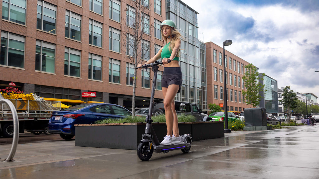 A woman riding the GOTRAX G5 commuter electric scooter on a wet sidewalk.