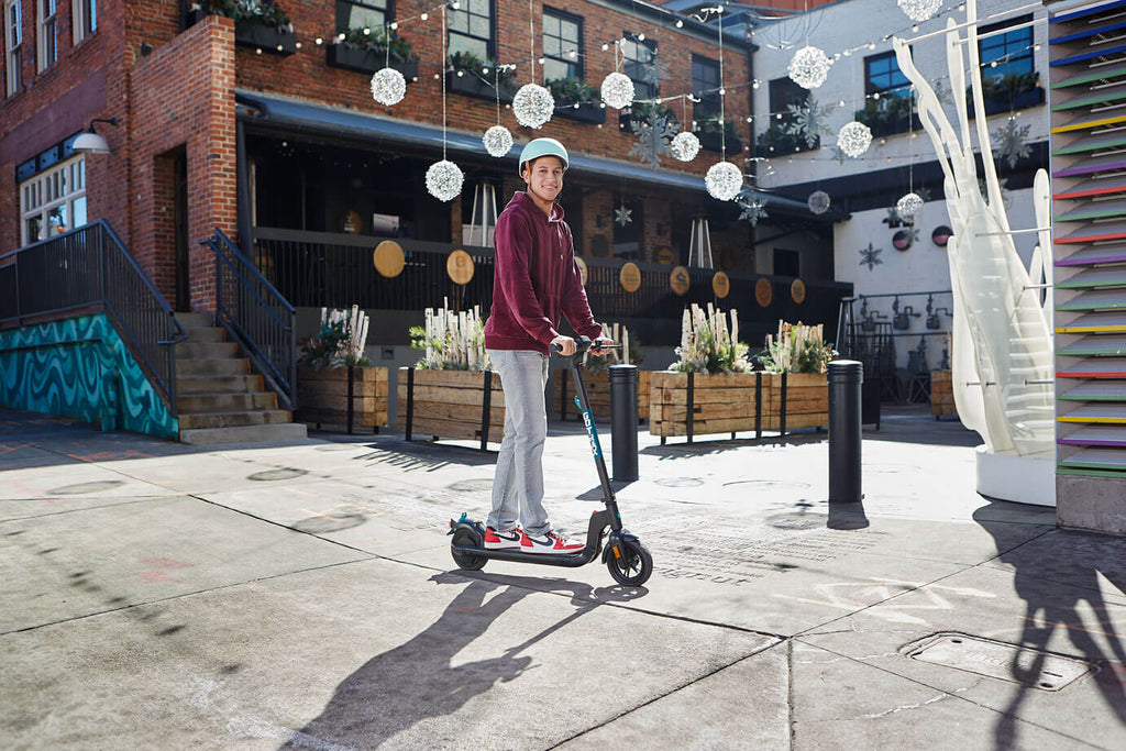 A person in a red hoodie standing on the GOTRAX Apex foldable electric commuter scooter with a rear-wheel motor.