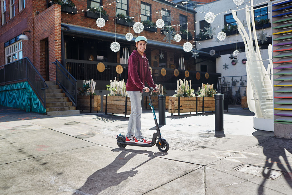 A man standing in front of winter decorations on a GOTRAX Apex Foldable Electric Commuter Scooter with a digital display and rear-wheel drive.