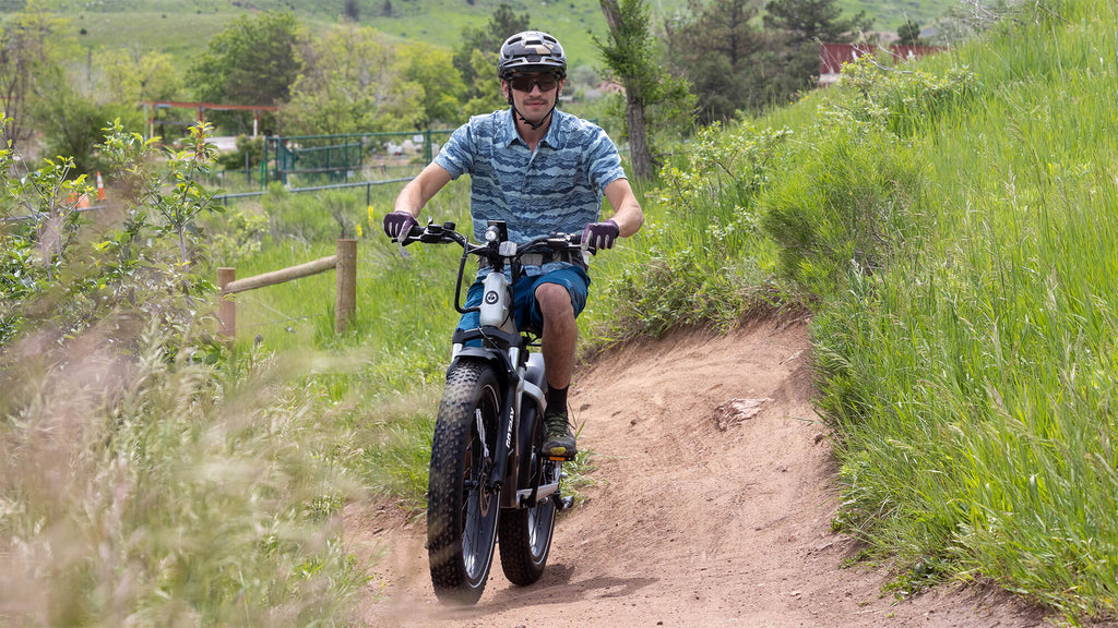 A man riding the GOTRAX Tundra off-road electric bike with fat tires and a torque sensor on a grassy hillside dirt path.