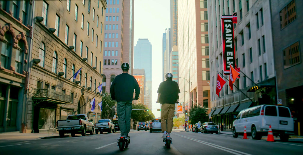 Two men riding GOTRAX GX1 and GX2 electric scooters with dual motors and dual suspension on a city street.