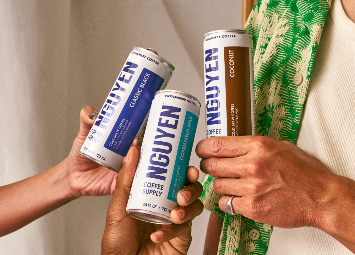 Three hands holding canned Vietnamese coffee drinks with branding visible.