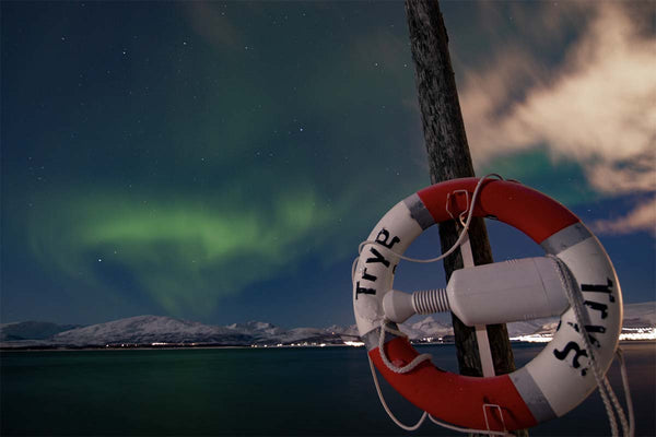 The aurora dances in the cloudy sky over a body of water with snow capped mountains in the distance. In the foreground a life buoy ring hangs from a wooden pillar