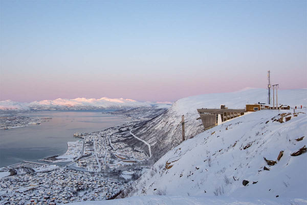 The sky is a gradient from blue to pink as the sun sets with snow capped mountains overlooking a small Norwegian town