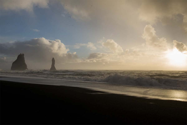 A black sand beach is illuminated by a setting sun in a cloudy blue sky. In the distance tall pillars of rock stand in the sea