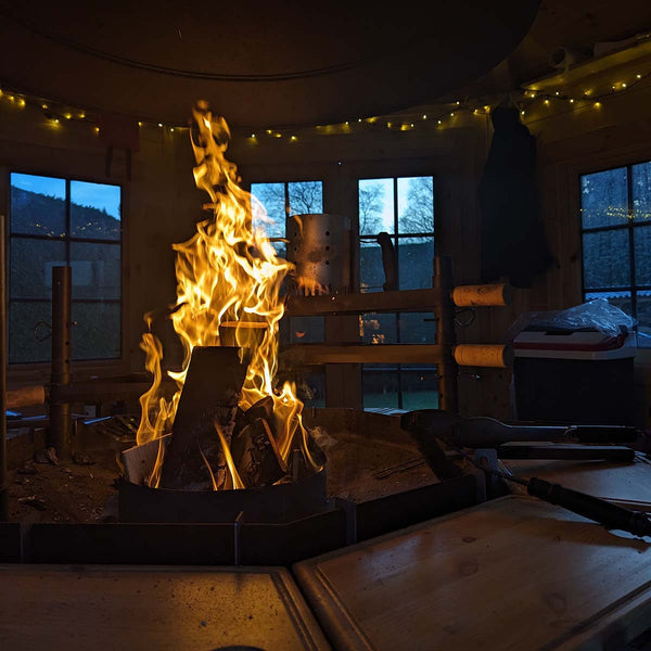 A large fire burns in the foreground in a small wooden cabin. In the background fairy lights twinkle while the large windows show it's getting dark outside