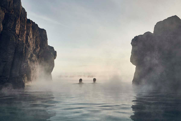 Two people in the distance are seen in the geothermic pool with steam rising from the water, man made rocks frame either side of the image