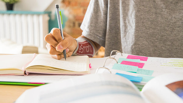 A young woman takes notes out of a notebook with sticky notes and organizational tools utilized; she has an eSmartr smart compression sleeve on her arm.