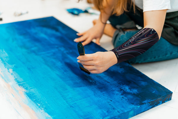 A woman paints blue on a canvas using a smart compression sleeve to help reduce cognitive stress.