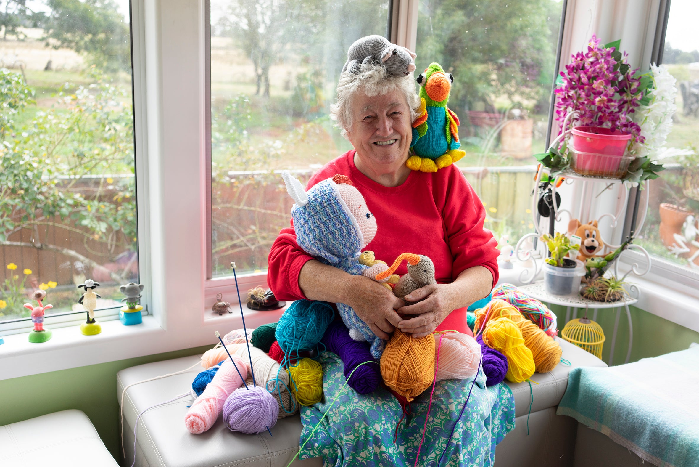 Tina Wilson's mum, Joan, sits by a window. She's smiling and is surrounded by her colourful knitted toys and creatures.