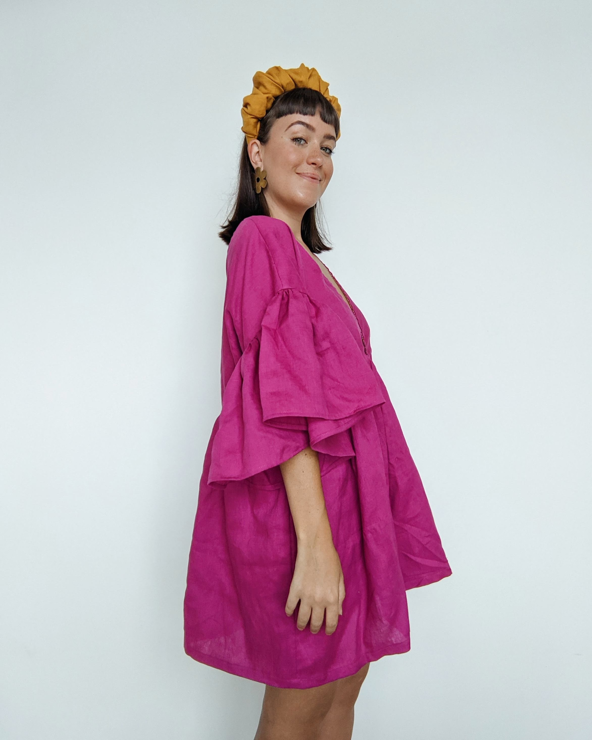 Daisy stands side on smiling towards the camera.  She is wearing a magenta pink linen dress with a golden linen headband.
