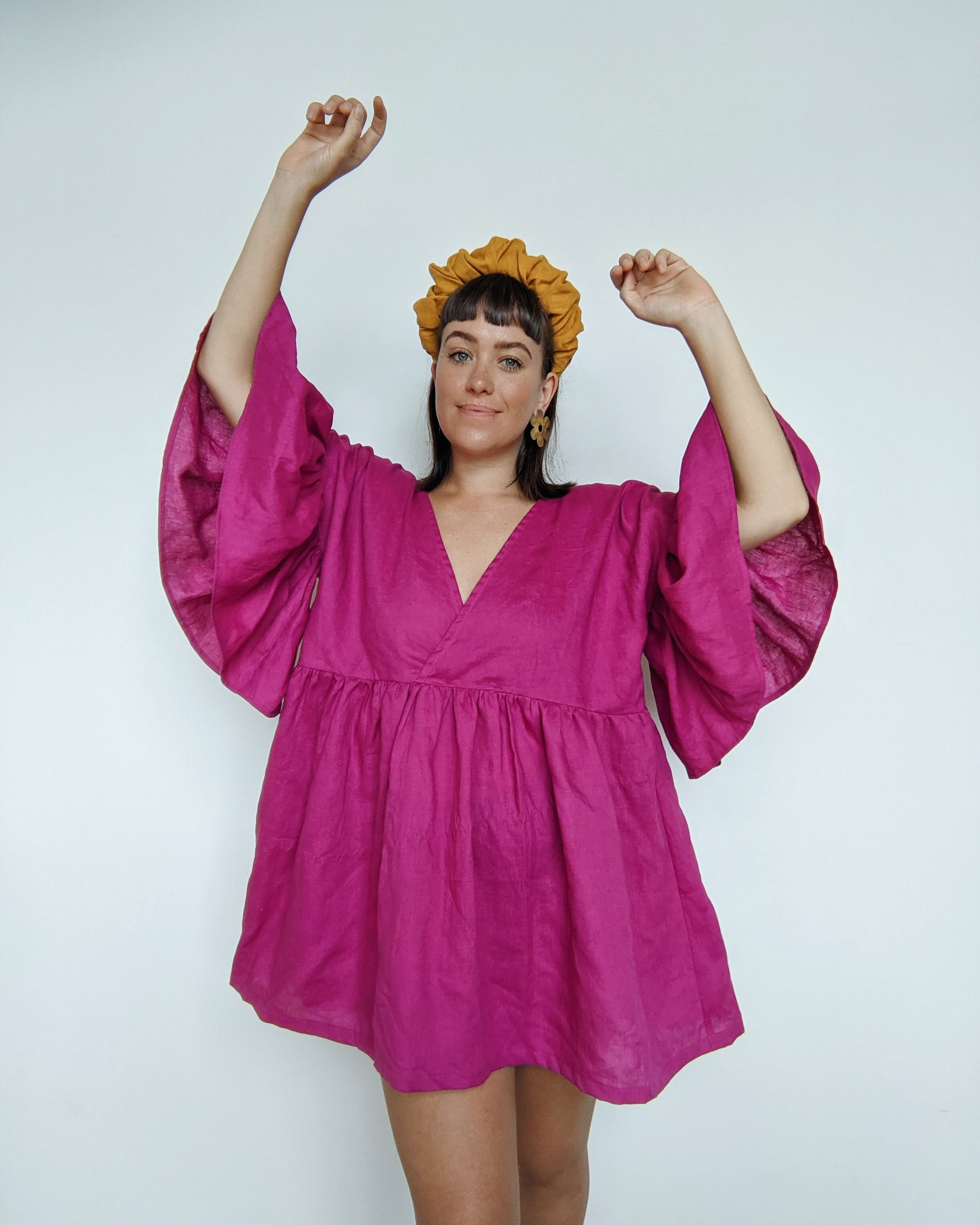 Daisy stands with her hands in the air to show the volume in the ruffle sleeves on her pink linen Maya ruffle dress hack.