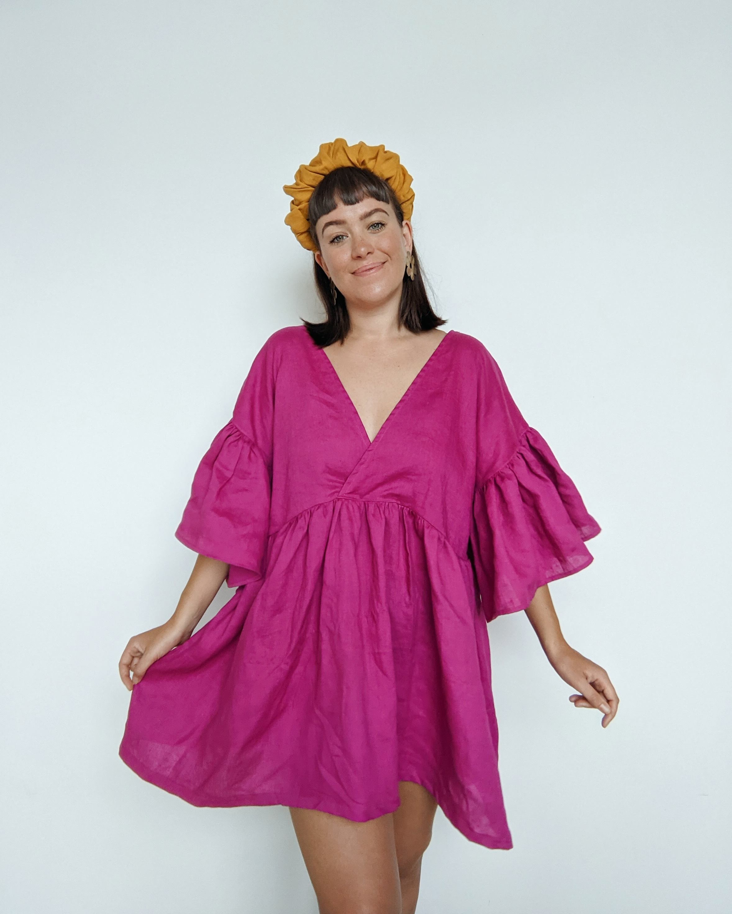 Daisy wears a vibrant, bright pink linen dress with a golden fabric crown headband.  She is holding one side out slightly to show the volume in the Maya ruffle dress skirt. 