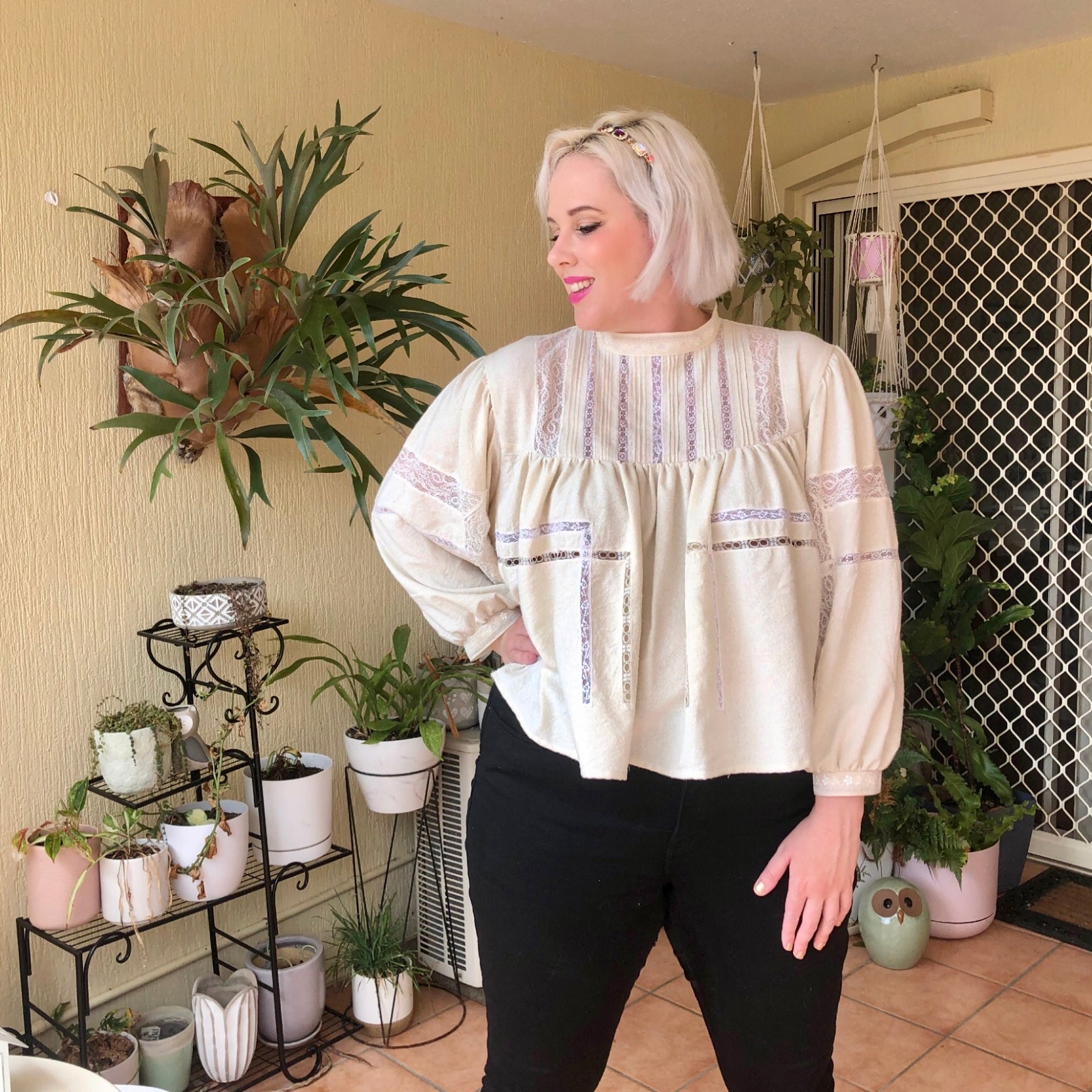 Shannon stands with one hand on her hip in front of her potted plants wearing her off white Silk Noil and lace long sleeved blouse with black jeans.  She is looking off to the side.