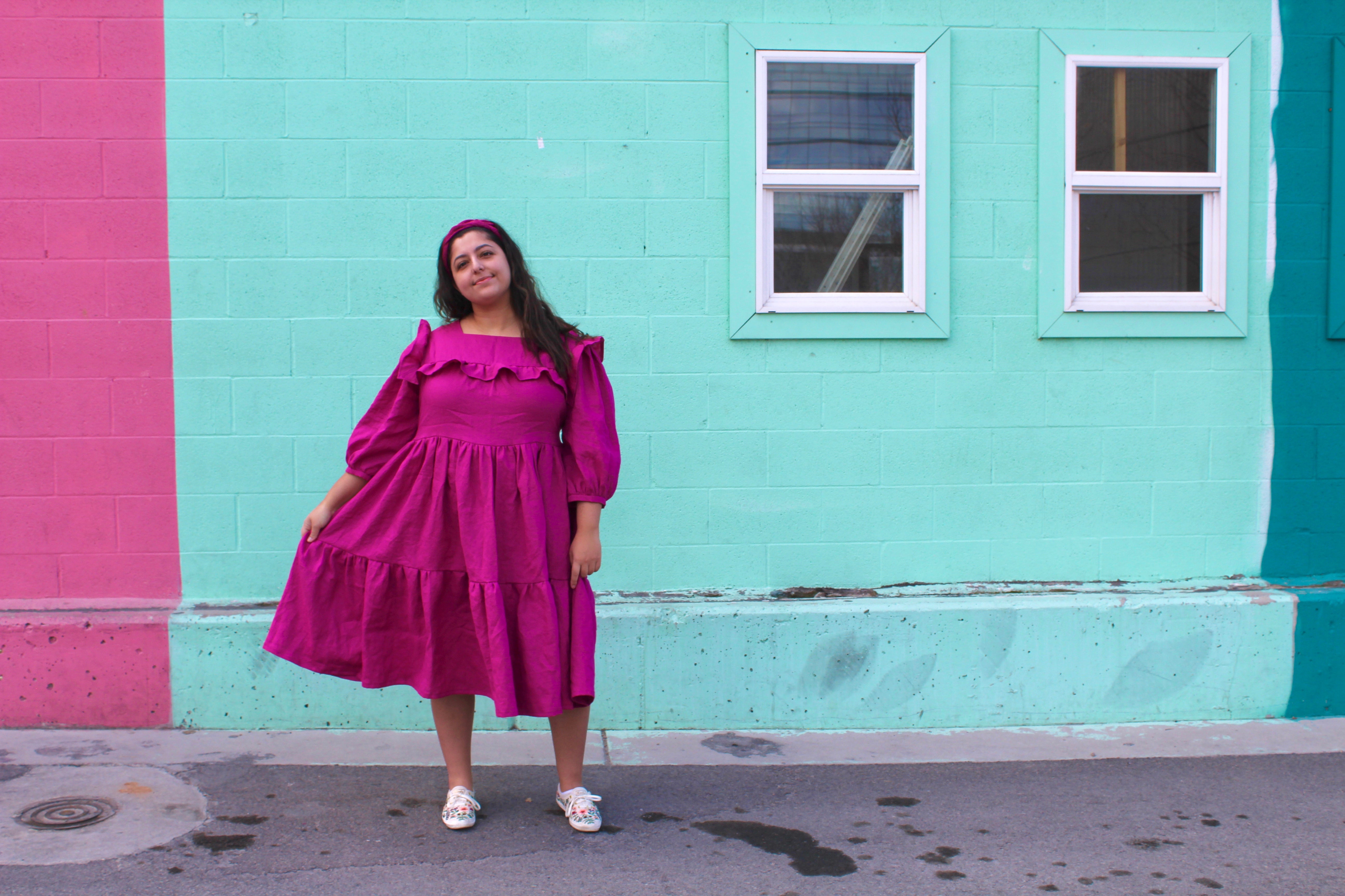 Romy wears a bright pink linen handmade dress, holding one side out slightly from her body to show the vlolume in the tiered skirt.  She is standing in front of a light blue, minty  painted wall.