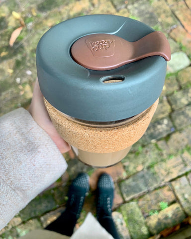 Downward view of woman in coat holding grey and cork keepcup over brick path