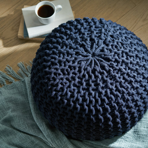 Navy Blue Knitted Round Pouf on top of a teal throw blanket next. A cup of coffee is perched on top of a stack of three books in the upper left.