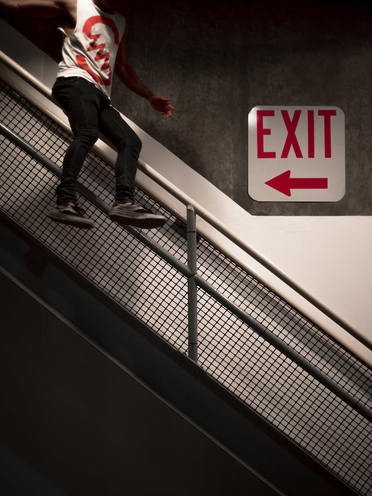 A man sliding down a handrail in a dimly lit stairwell. His shirt is white with a red conductor symbol and it matches the EXIT sign seen hanging on the wall.