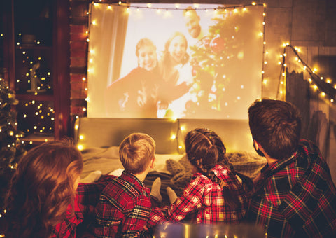 New Year's Movie Night. A family with 1 father. 1 mother,1 little girl and 1 little boy are watching a movie together in their pyjamas. They watch it on a big white screen with decorative lights around. It's seems very cozy and relaxing.