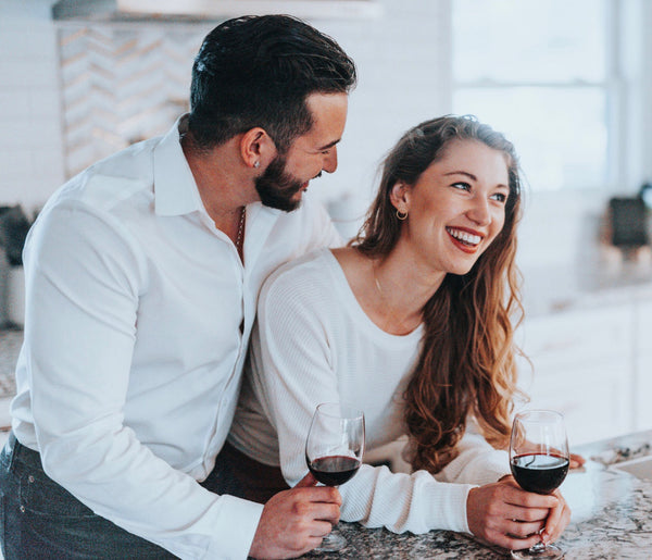 10 Ways on How To Be a Better Lover: Couple smiling as they enjoy wine together in the kitchen.