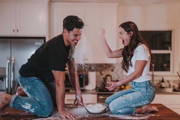 How To Be a Better Lover: Couple sharing laughs as they throw flour at each other on kitchen counter.