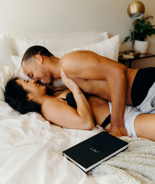 10 Ways on How To Be a Better Lover: Couple smiling and leaning in for a kiss as they lie in bed with The Adventure Challenge Book beside them.
