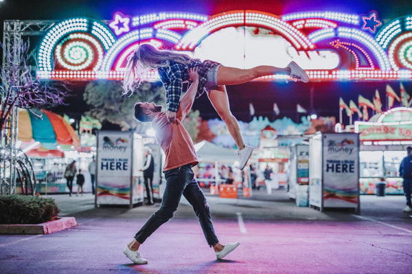 An image of a boyfriend lifting his girlfriend up over his head while they are at a carnival together.