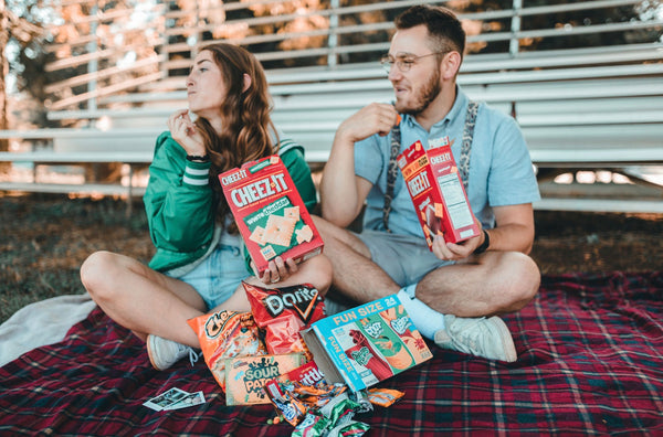 A couple sit on a picnic blanket and eat nostalgic snacks together.
