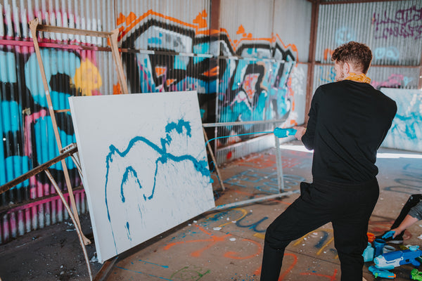 A man uses a squirt gun to do squirt gun painting against a canvas in a warehouse.