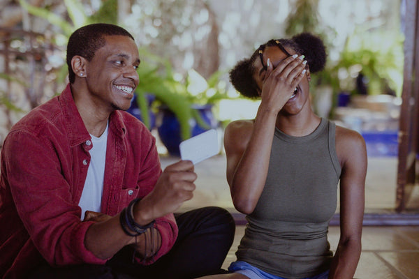 Husband smiles as he holds a card in his hands while his wife laughs and puts her hand on her face in embarrassment