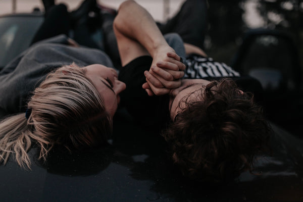 Husband holds his wife's hand and plants a kiss on it as they lie on the hood of their car