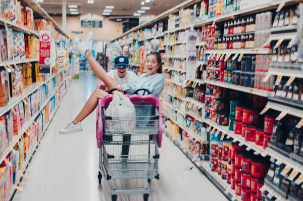 Two best friends crushing their best friend goals and having fun at the grocery store, with the girl sitting in the push cart and the guy pushing her across the aisle