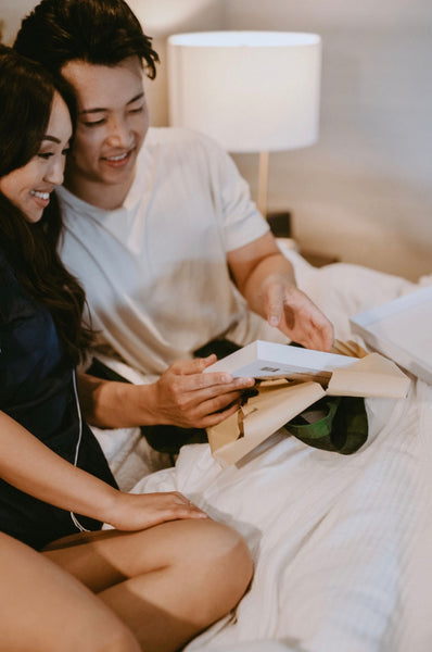 Creative, Romantic, and Fun Stay-at-Home Date Ideas for Couples: Couple smiling as they open a gift on the bed