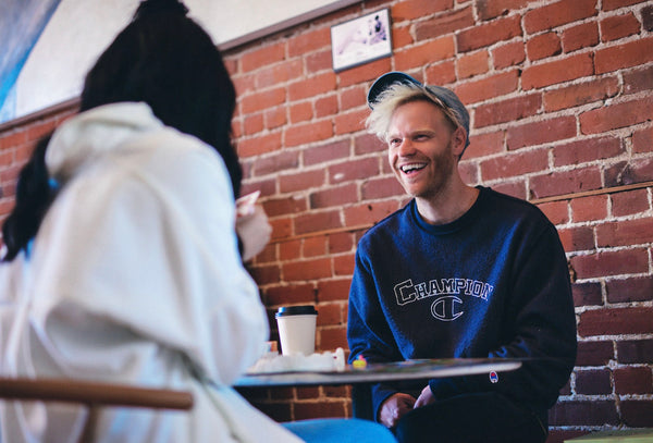 A girl with her back to the camera talking to a guy who is laughing at her jokes and maintaining good eye contact with her while sitting on a cafe table and enjoying their coffee