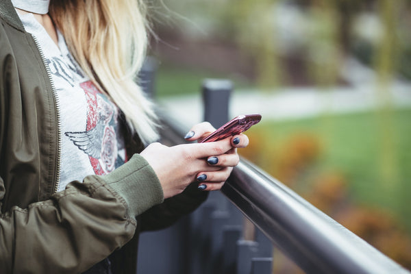 ​​How to Get the Best Deals on Back to School Supplies. Close-up photo of girl wearing a green jacket using her phone next to gray railings.