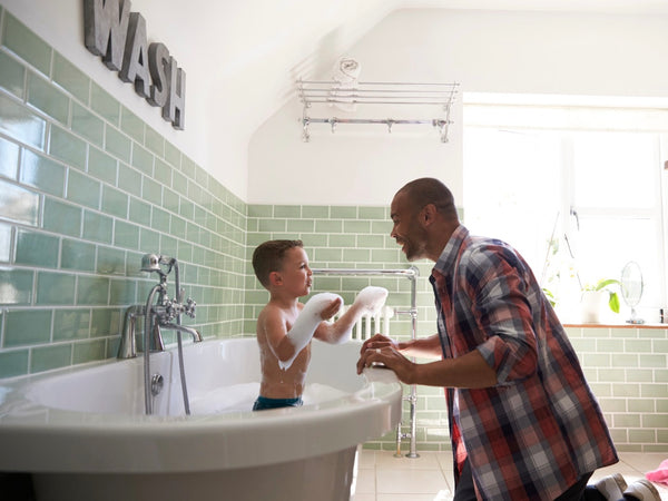 How to get kids to sleep better: Dad smiles and plays with his son as the latter enjoys a bubble bath before bed.