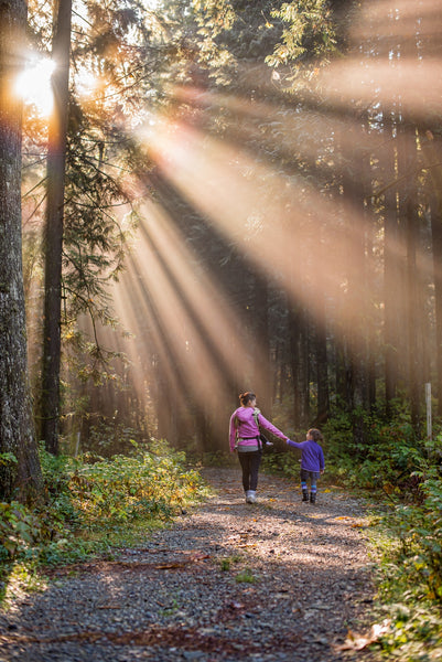 A mom and her son walk on a trail in the woods while the sun is shining through the trees.