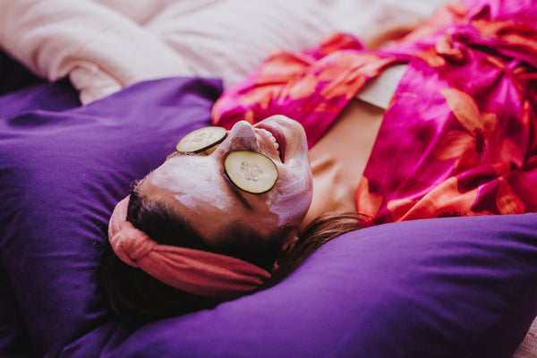 An image of a woman lying on a bed at a spa with cucumbers on her eyes and face mask on.