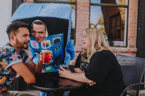 A girl and guy sit at a table chatting while a man dressed in an iPhone costume whispers in the males ear