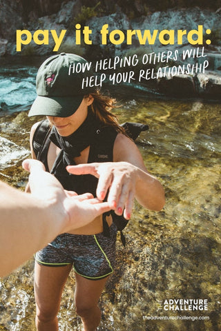 A man holds out his hand for his girlfriend to grab as they walk up the side of a cliff on a hike; image overlaid with text that reads pay it forward: how helping others will help your relationship